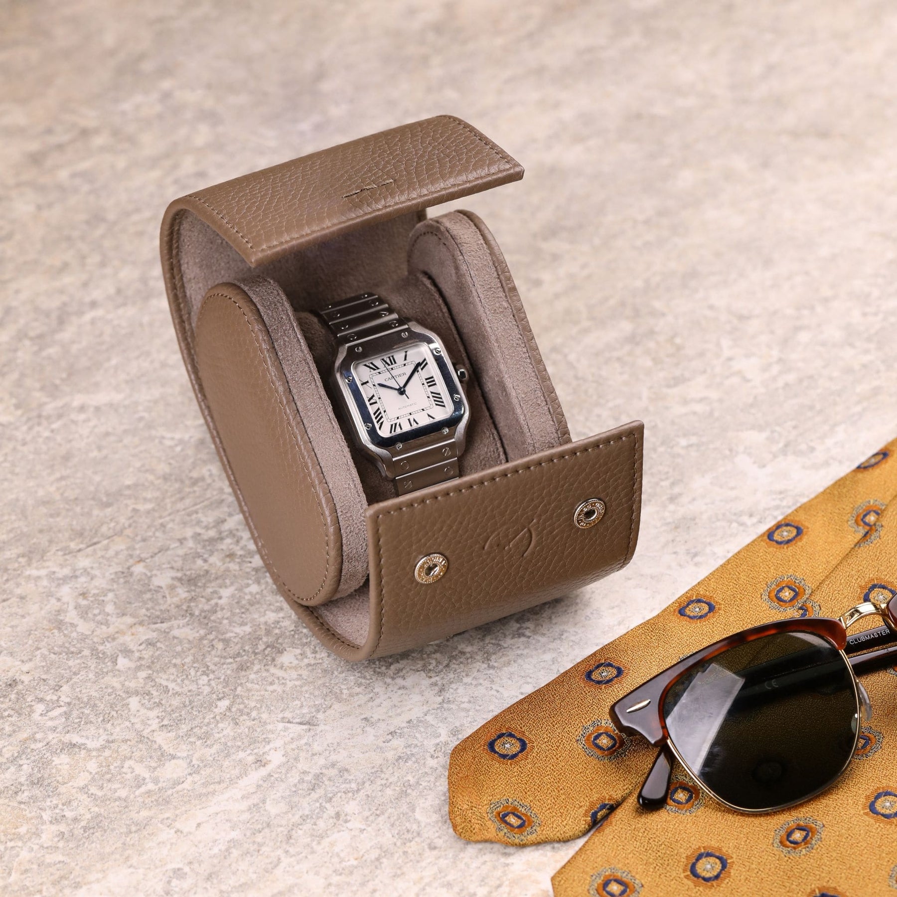 roll porta orologio in pelle color tortora con Cartier Santos all'interno e occhiali da sole e cravatta vintage sull'angolo destro della foto
