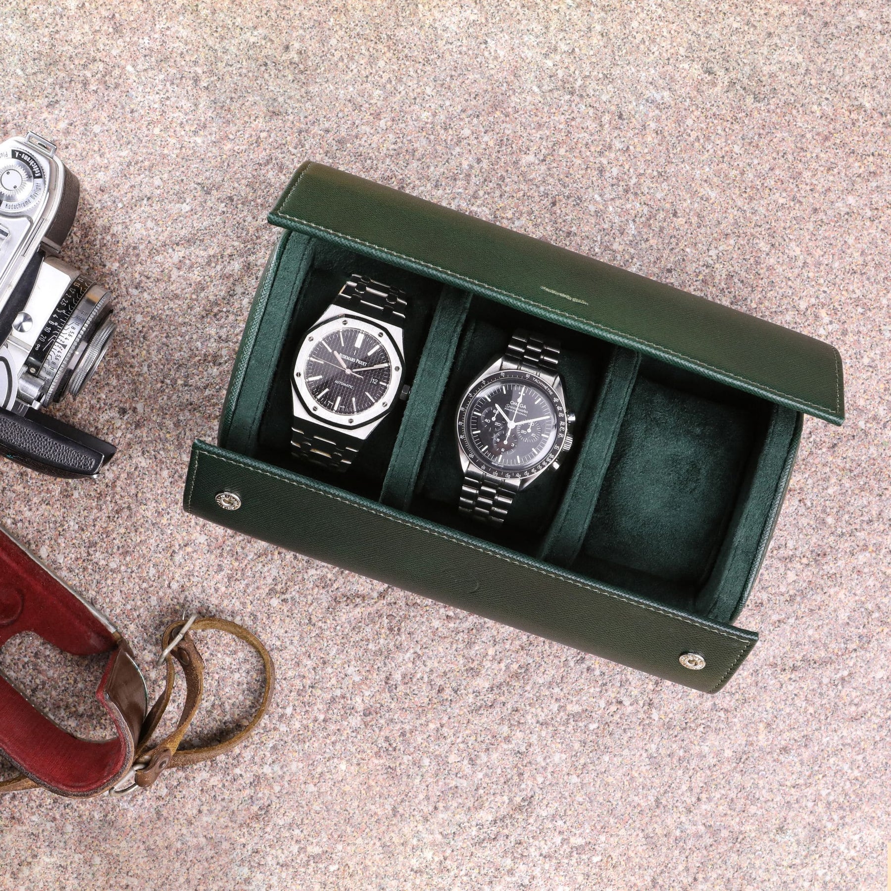 Rotolo porta orologi da 3 in pelle saffiano verde con Audemars Piguet Royal Oak e Omega Speedmaster Moonwatch all'interno e macchina fotografica vintage al lato sinistro